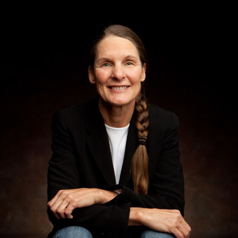 The image shows a woman with long brown hair styled in a braid. She is wearing a black blazer over a white t-shirt and blue jeans. She is smiling and looking directly at the camera. The background is a dark brown color.